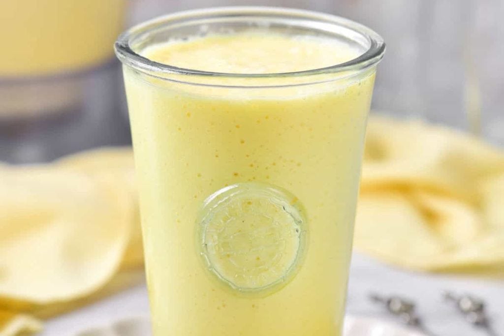 A glass filled with a yellow smoothie on a white plate.