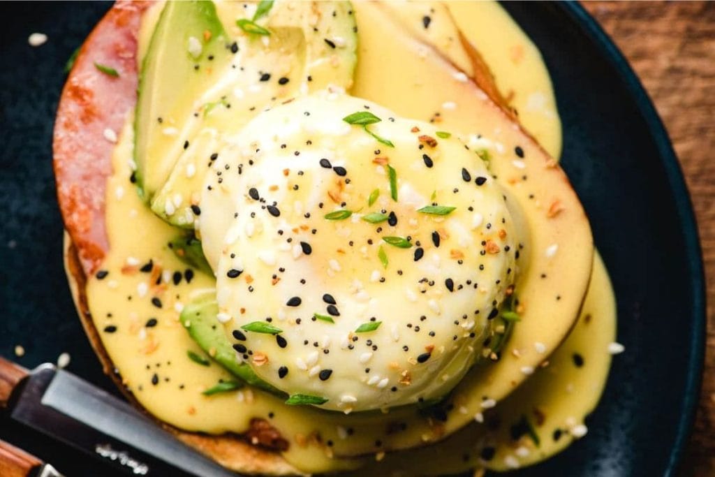 Eggs benedict with avocado and sesame seeds on a black plate.