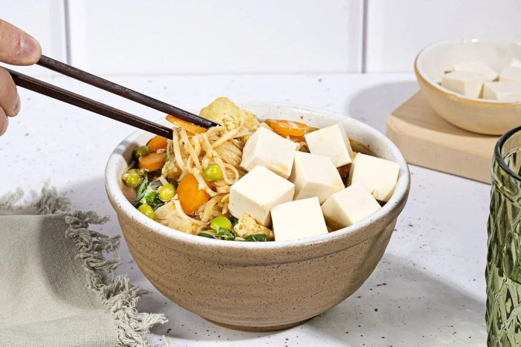 A person is holding chopsticks over a bowl of noodles and tofu.