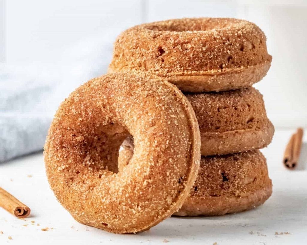 A stack of healthy, cinnamon sugar-coated doughnuts on a light background with cinnamon sticks nearby.