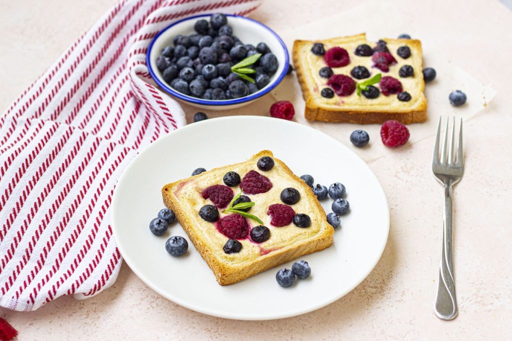 Two slices of bread topped with peanut butter and assorted berries on a white plate, with extra blueberries in a small bowl and a red-striped towel on the side, serving as healthy snacks.