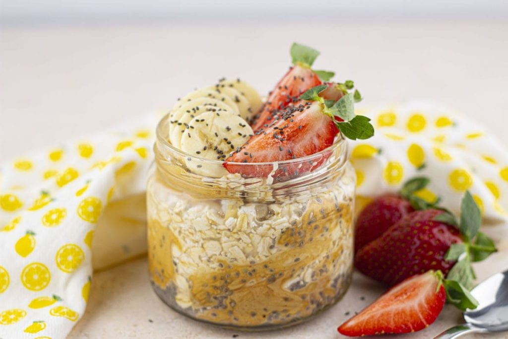 A jar of overnight oats topped with banana, strawberries, and chia seeds, accompanied by fresh strawberries on a table with a yellow floral cloth, ready to be enjoyed as healthy snacks.