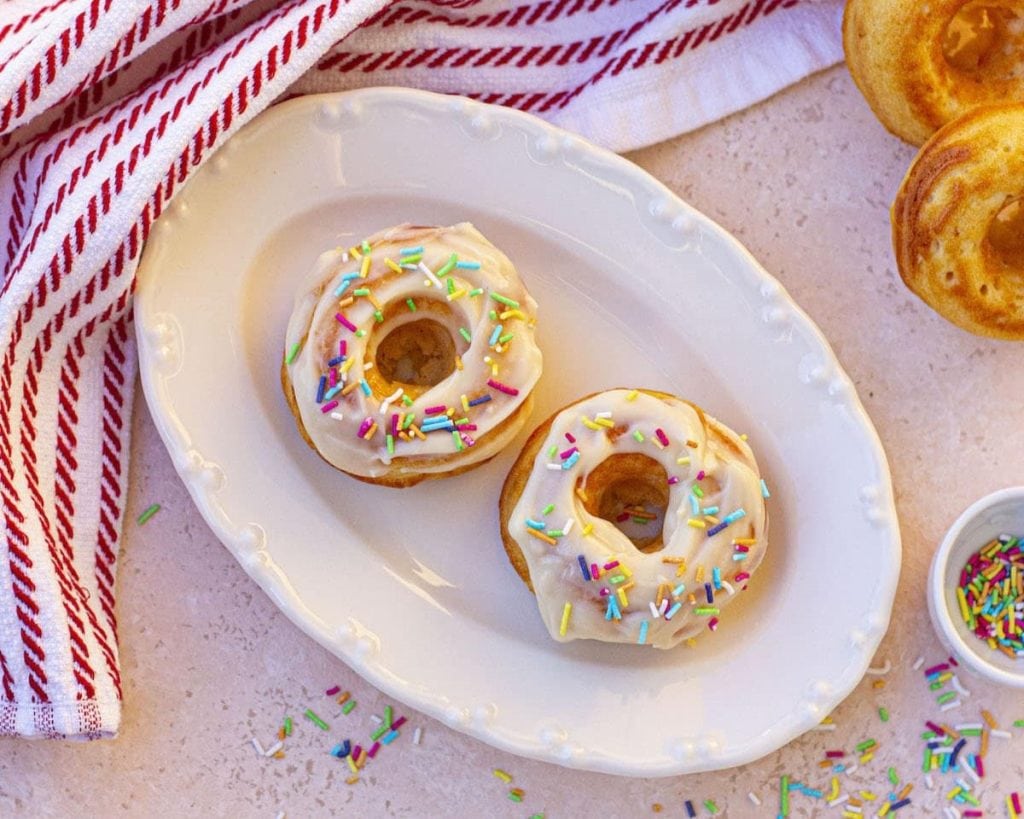 Two healthy frosted doughnuts with sprinkles on a white plate, accompanied by a striped napkin and extra sprinkles on the side.