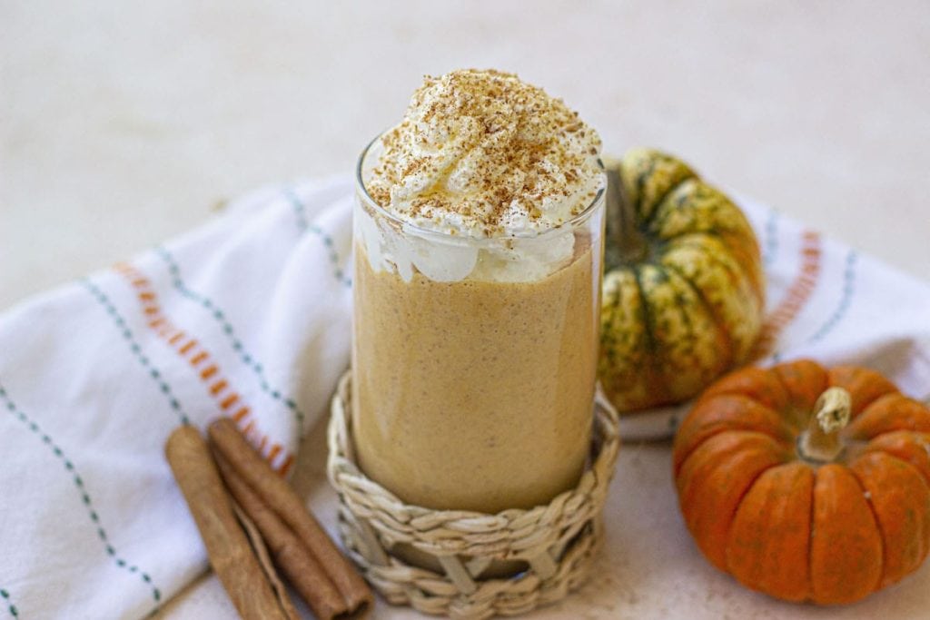 A glass of pumpkin spice latte topped with whipped cream and cinnamon, accompanied by healthy snacks, mini pumpkins, and cinnamon sticks.
