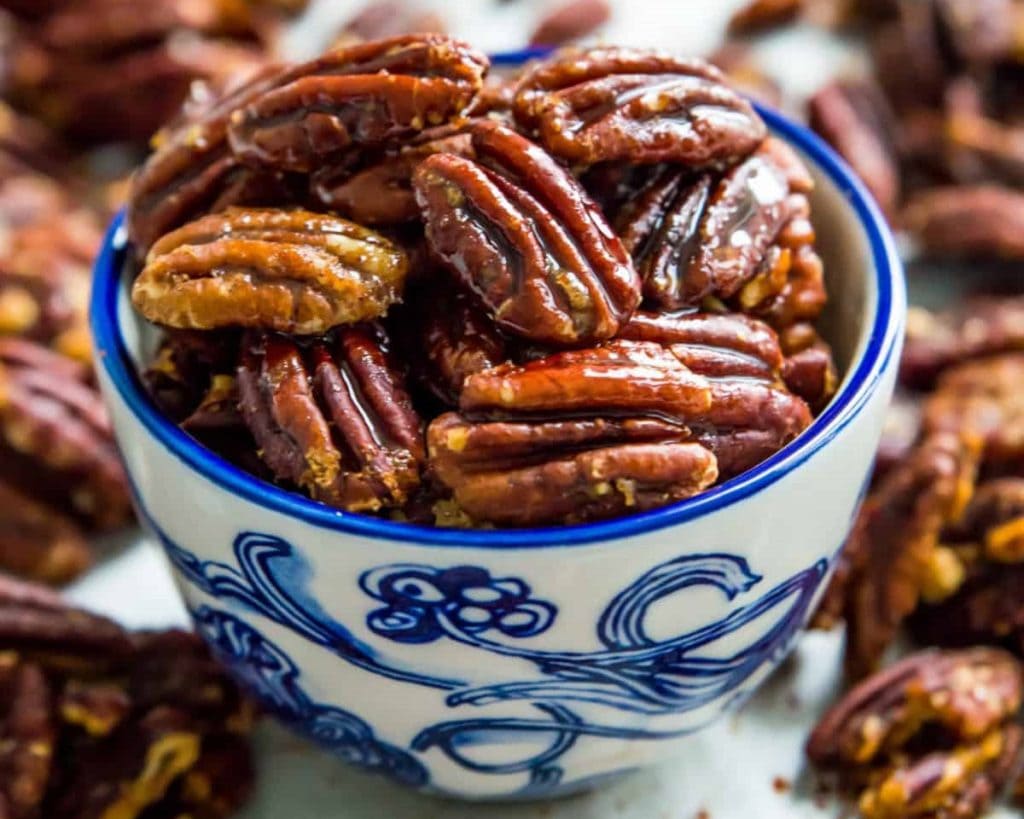 A bowl of glossy pecan nuts, a healthy snack, on a surface surrounded by more pecans.