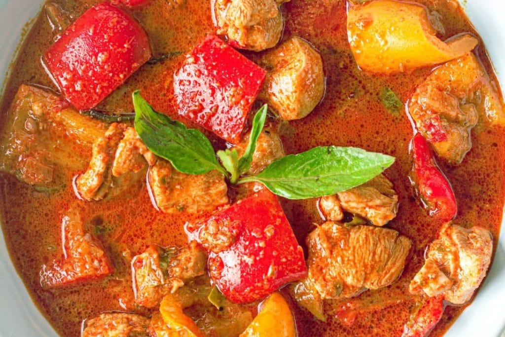Close-up image of thai red curry, perfect for date night dinners, with chunks of meat, bell peppers, and thai basil leaves in a rich, spicy sauce.