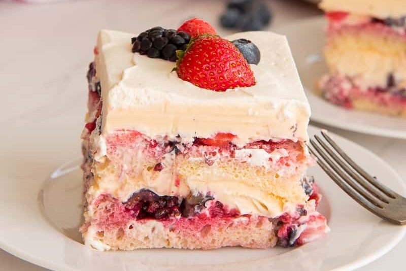 A slice of layered dessert with cream, sponge cake, fruit, and topped with strawberries and blackberries, served on a white plate with a fork on the side. One of the best summer cakes to enjoy!