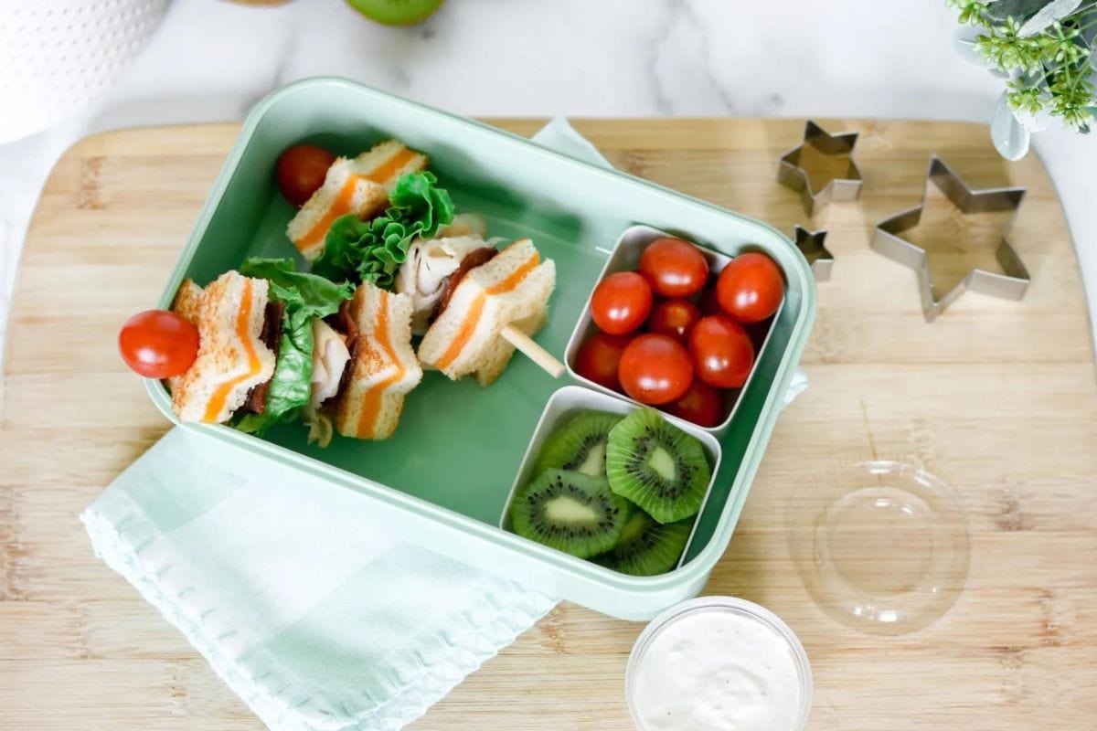 A lunch box with a sandwich skewer topped with tomatoes, sections of cherry tomatoes, and kiwi slices, placed on a wooden board with star-shaped cookie cutters nearby.