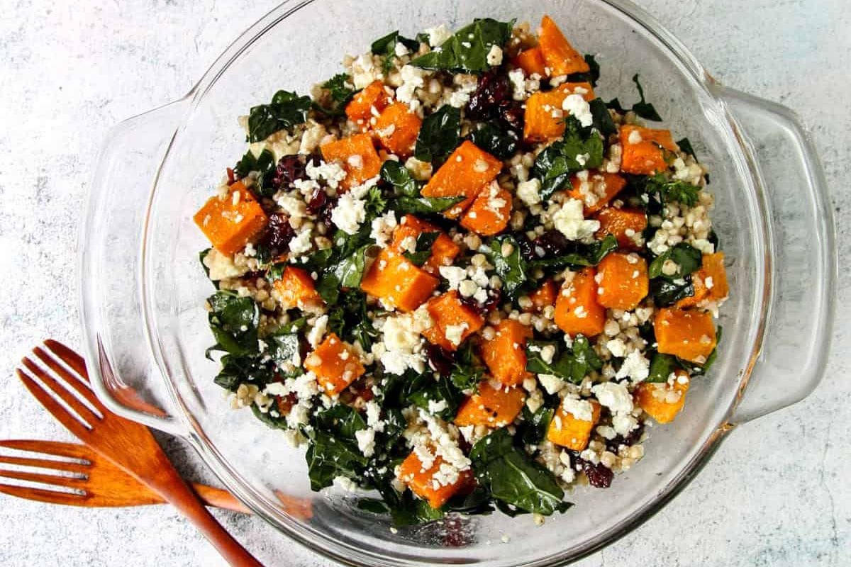 A glass bowl filled with a kale salad featuring roasted sweet potatoes, couscous, crumbled cheese, and dried cranberries, with wooden utensils beside it.
