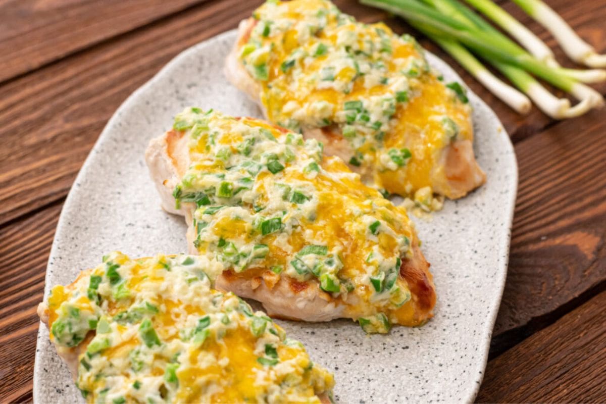 Three pieces of baked chicken breast topped with melted cheese and chopped green onions, adding a zesty twist to classic jalapeno recipes, on a speckled white plate, placed on a wooden table. Green onions are visible in the background.