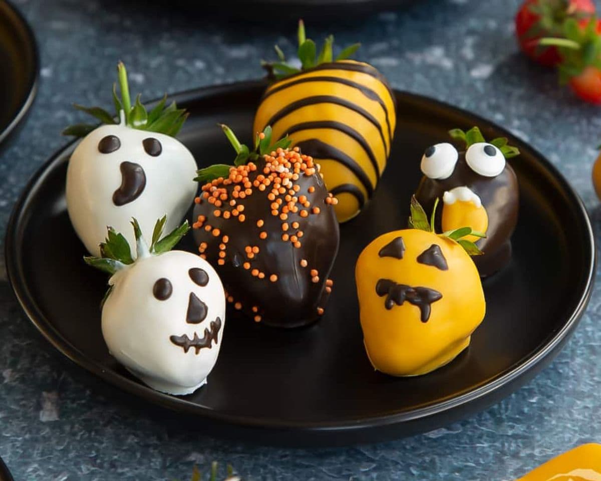 A black plate holds six strawberries dipped in chocolate, decorated with Halloween-themed designs including ghosts, a pumpkin, a mummy, and a beehive—perfect examples of easy Halloween foods.