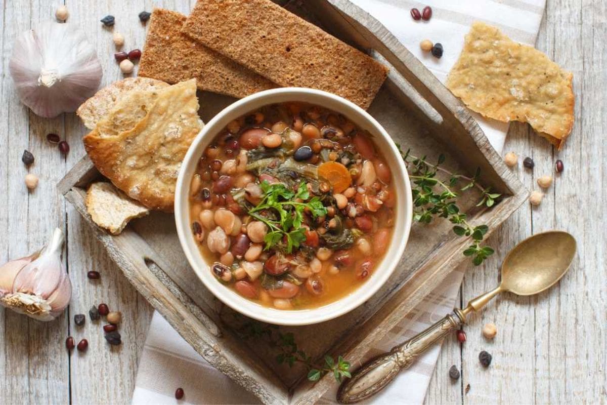 A wooden tray holds a bowl of bean soup garnished with greens, flanked by various types of bread, garlic cloves, and a spoon. Dried beans and thyme sprigs are scattered around the tray.