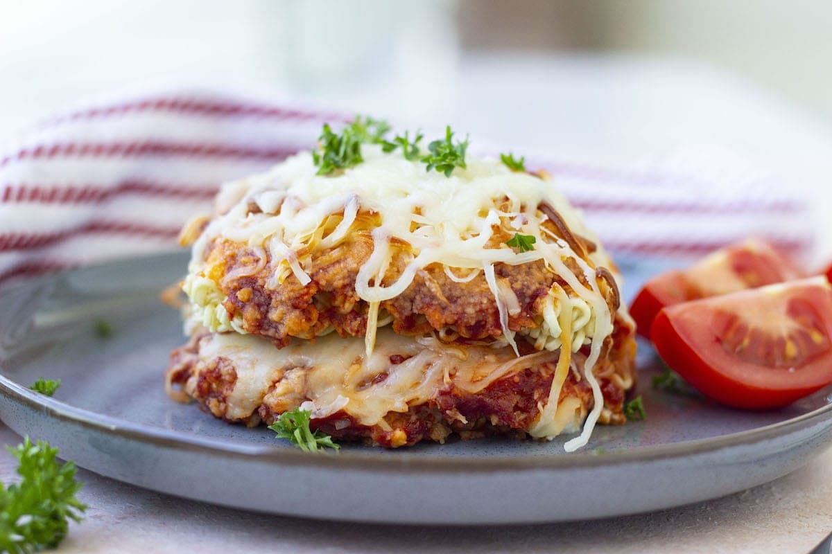 A plate with a serving of lasagna topped with melted cheese, garnished with parsley, and accompanied by sliced tomatoes, making it one of the perfect Christmas dinner ideas.