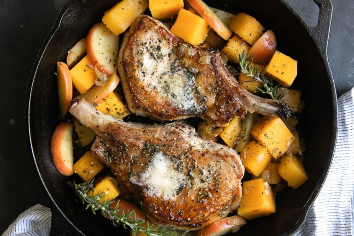 A cast iron pan containing two cooked pork chops seasoned with herbs, surrounded by diced butternut squash, apple slices, and sprigs of rosemary.