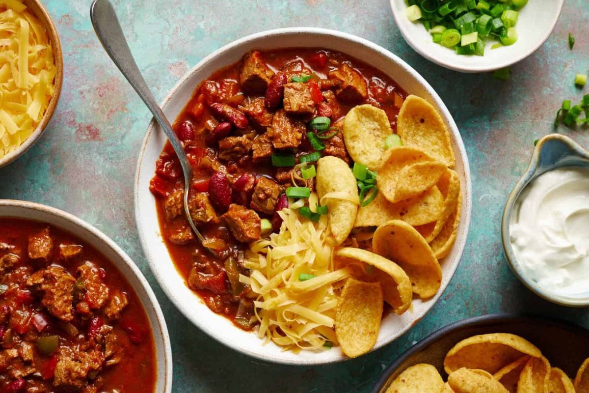 A bowl of chili with meat and beans is garnished with shredded cheese, green onions, and corn chips. It is accompanied by bowls of shredded cheese, sliced green onions, and sour cream.