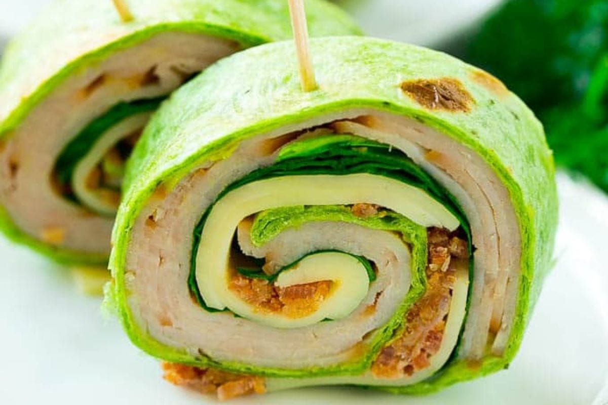 Two pinwheel sandwiches made with green spinach tortillas, sliced turkey, cheese, lettuce, and bacon, held together with toothpicks—perfect lunch ideas for school.