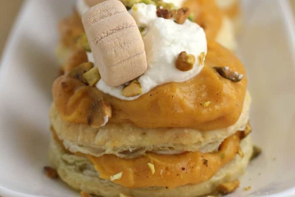 Close-up of a dessert with layers of pastry and pumpkin-colored cream, topped with chopped nuts, a dollop of white cream, and a rectangular biscuit on a white plate—a perfect fall recipe dessert.