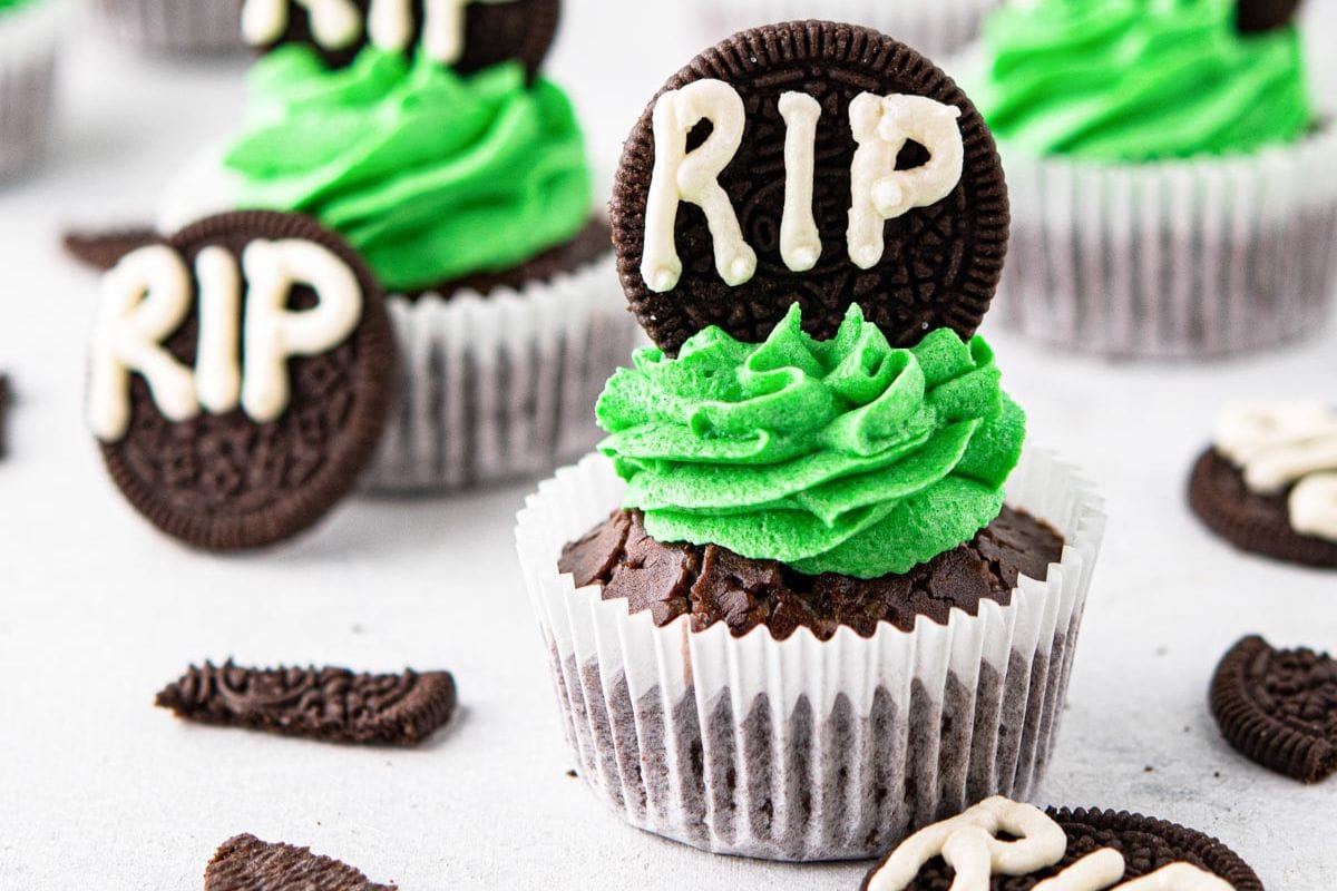 Halloween cupcakes ideas come to life with these chocolate cupcakes, adorned with green frosting and decorated with cookies inscribed "RIP" in white icing, all arranged on a white surface.