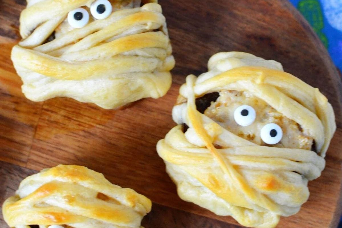 Three pastry-covered treats resembling mummies with googly eyes, placed on a wooden surface.