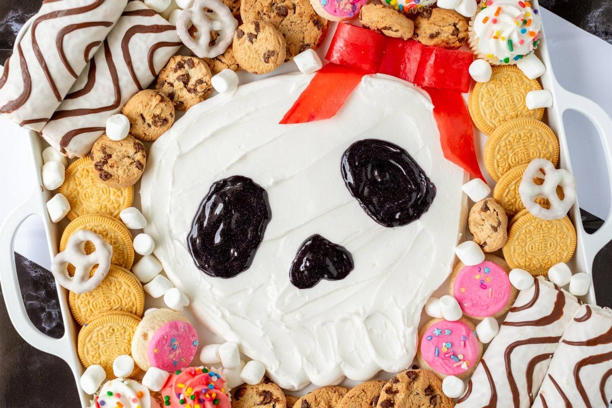 A Halloween-themed dessert platter with cookies and a skull design made from frosting and chocolate. The skull has red candy for a bow and is surrounded by colorful cookies and marshmallows.