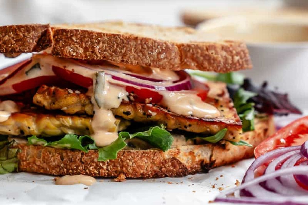 Close-up of a sandwich with grilled tofu, lettuce, tomato, red onion, and sauce between two slices of whole grain bread. This nutritious option fits perfectly among middle school lunch ideas. Sliced red onion and tomato are visible in the foreground.