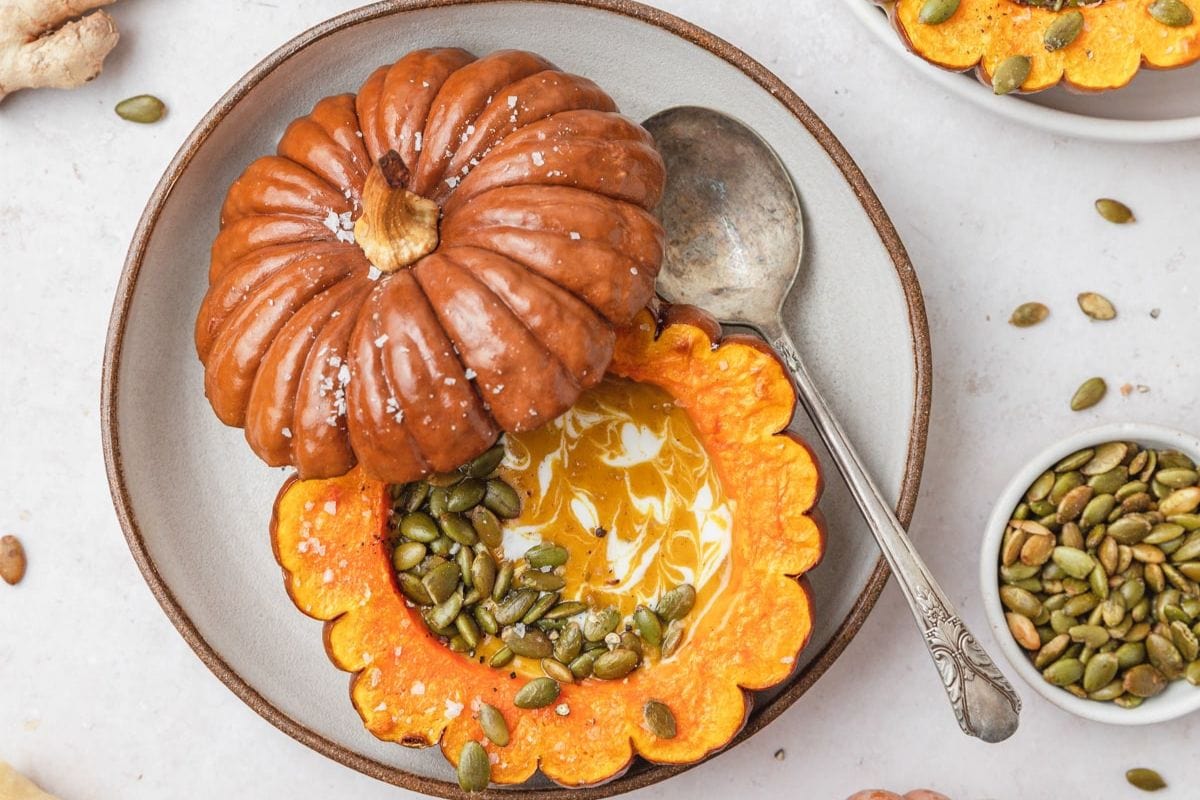 A halved stuffed pumpkin filled with soup, garnished with seeds, and served with a spoon on a grey plate.