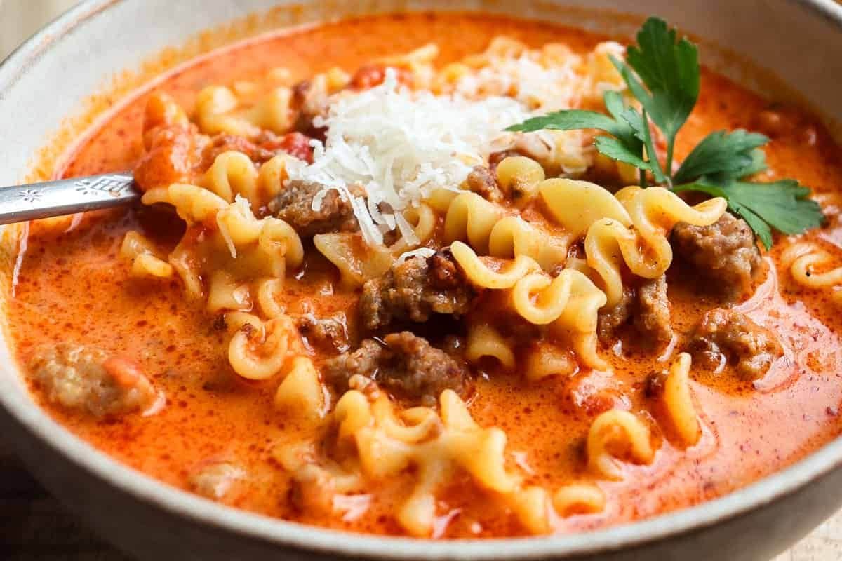 A bowl of creamy lasagna soup topped with shredded cheese and parsley, with a spoon placed on the side.