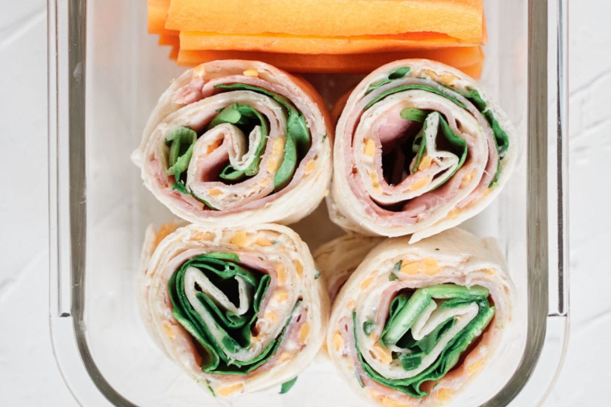         School Lunch Ideas: Lunchbox containing four tortilla pinwheels with ham, cheese, and spinach, and carrot sticks in a separate compartment.