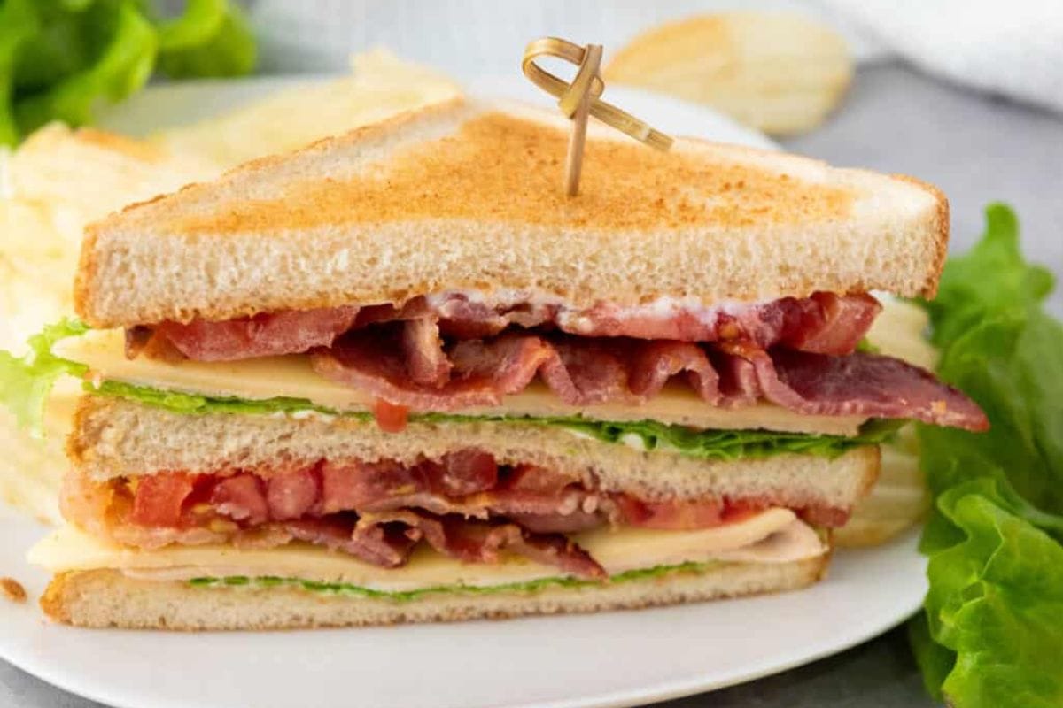 A close-up of a BLT sandwich, featuring toasted bread, layers of bacon, lettuce, tomato, and cheese, secured with a toothpick. Perfect for school lunch ideas for high schoolers, the sandwich is served on a plate with leafy greens and potato chips.