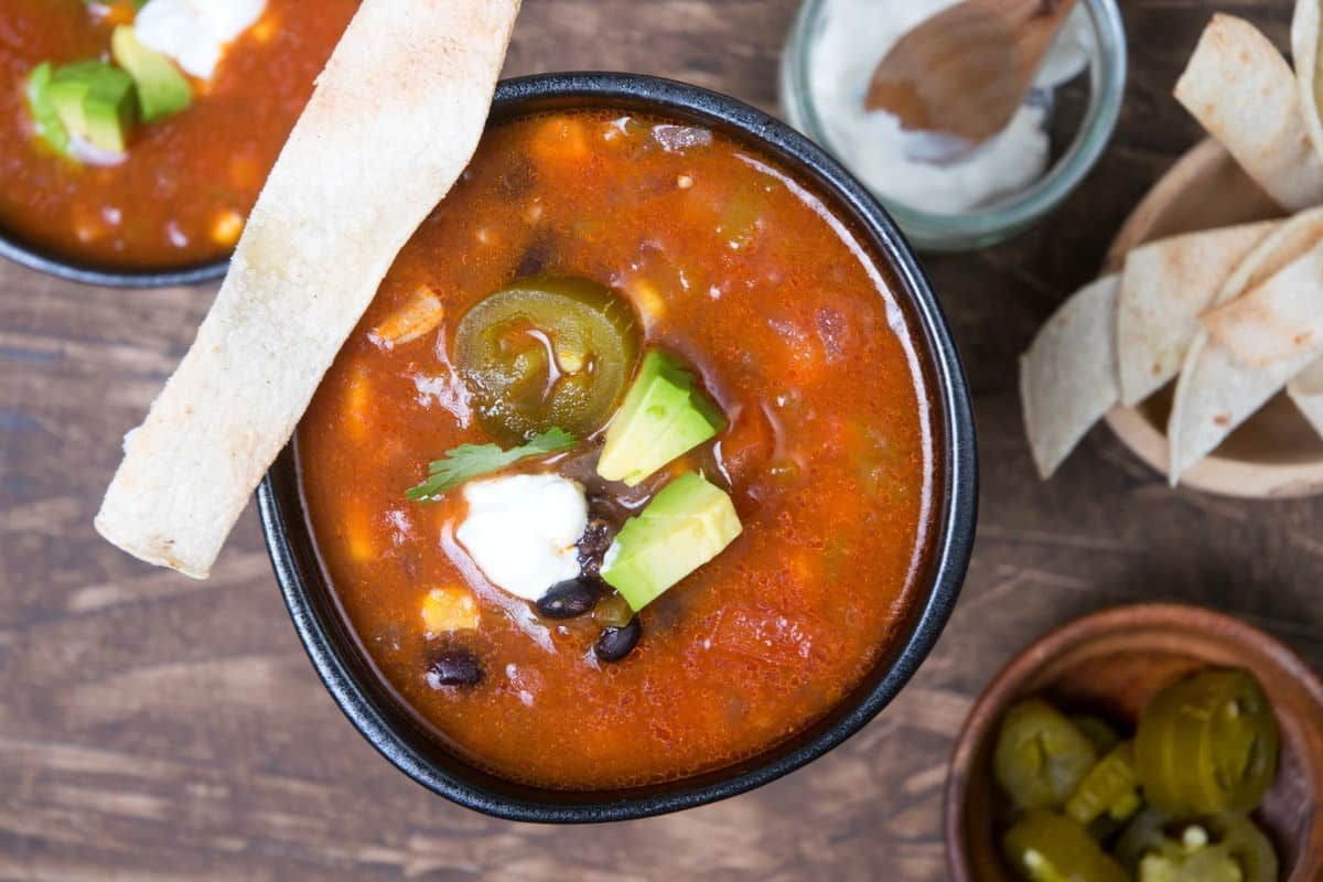 A bowl of soup garnished with avocado, jalapeno, sour cream, and tortilla strips. Nearby are a bowl of sliced jalapenos, a jar with a spoon, and a bowl of tortilla strips. Perfect for those who love Soup Crockpot Recipes.