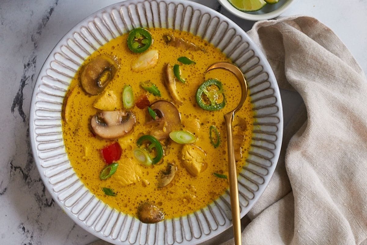 A bowl of yellow curry soup containing chicken pieces, sliced mushrooms, and green chilies with a spoon. A linen napkin is on the side, and a small bowl with lime wedges is in the background. Perfect for those who love discovering new soup recipes made in a slow cooker.