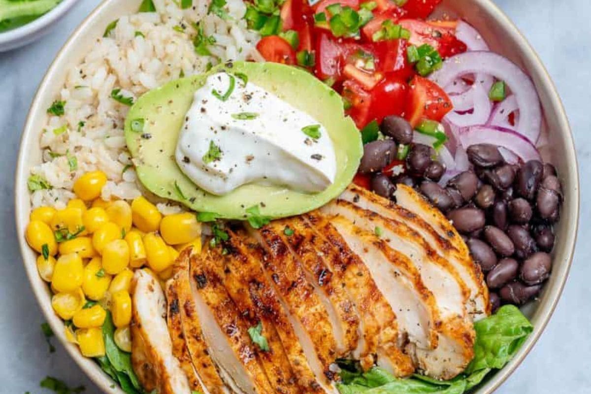 A bowl containing grilled chicken slices, avocado with sour cream, black beans, corn, brown rice, cherry tomatoes, and red onions, garnished with chopped herbs—perfect for quick and nutritious teacher lunch ideas.