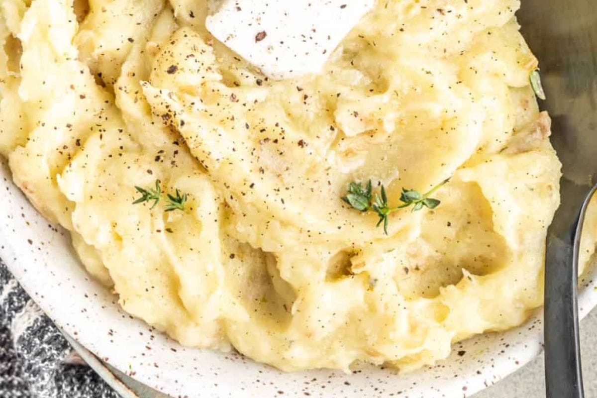 A close-up of creamy mashed potatoes, a quintessential Thanksgiving side dish, garnished with a sprinkle of black pepper, herbs, and a pat of butter in a speckled white bowl.