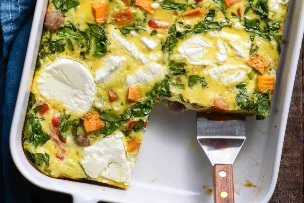Close-up of a baked vegetable frittata in a rectangular pan with a metal spatula lifting a piece. The dish includes visible ingredients like spinach, sweet potatoes, and sliced rounds of cheese.