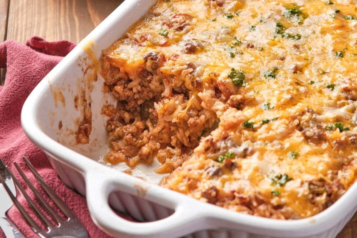 A white casserole dish with a portion of cheesy baked casserole with rice and ground meat missing. A red cloth and fork are nearby.