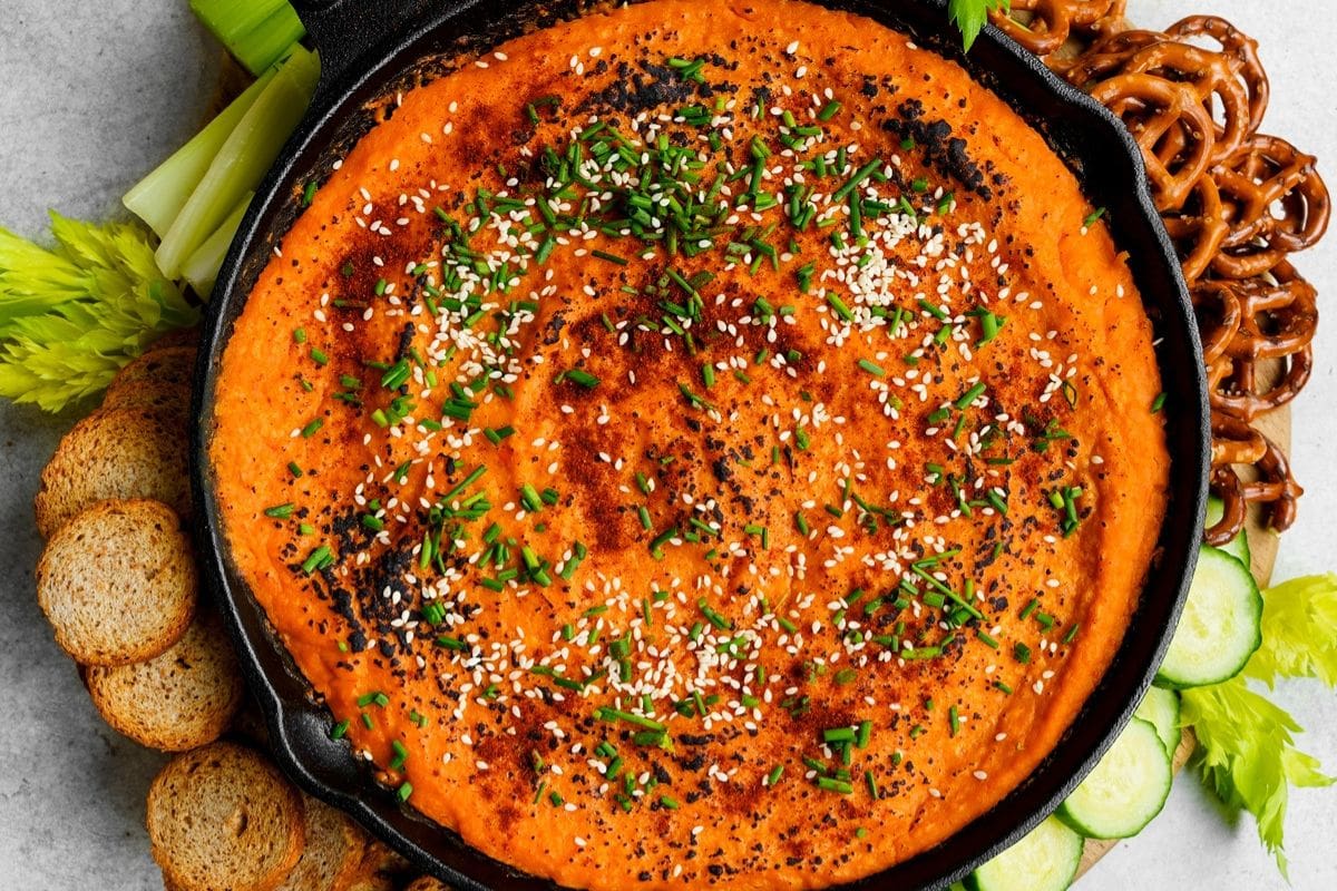 A skillet filled with creamy buffalo dip, garnished with sesame seeds and chives, surrounded by celery, sliced bread, pretzels, and cucumber slices. Perfect next to your favorite butternut squash recipes for a delightful fall spread.
