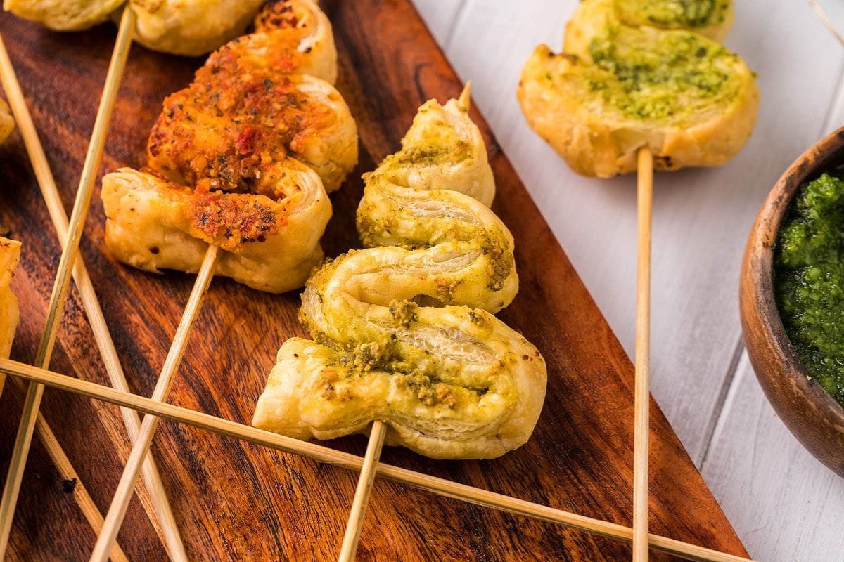 Puff pastry twists with pesto and tomato topping on skewers, placed on a wooden board, make delightful Christmas appetizer recipes. A bowl of green sauce is on the side.