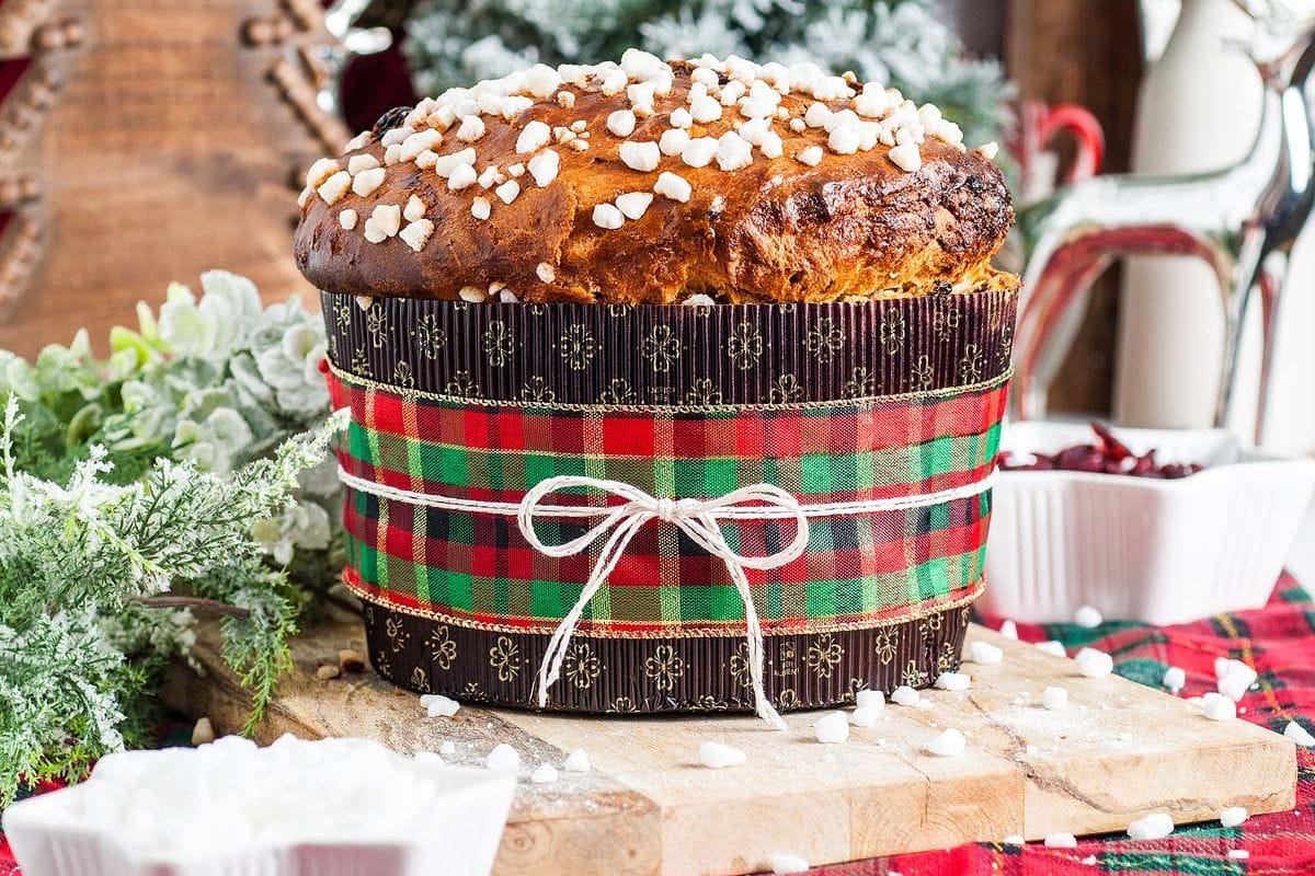 A traditional panettone, a classic in Christmas bread recipes, is adorned with sugar crystals and wrapped with a plaid ribbon, resting on a wooden board. Holiday decorations and greenery enhance the festive background.