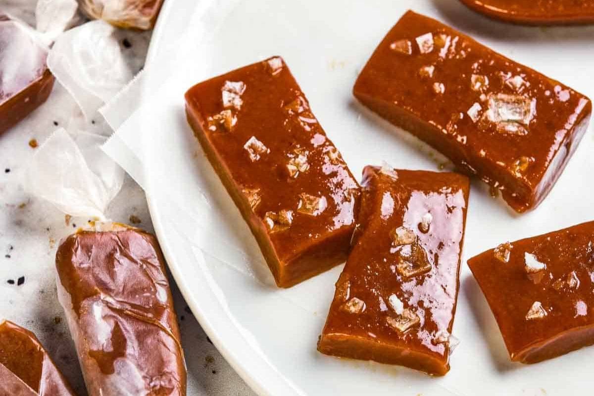 Glossy caramel candies with sea salt flakes are displayed on a white plate, evoking the charm of Christmas candy recipes, with some wrapped in wax paper to the side.