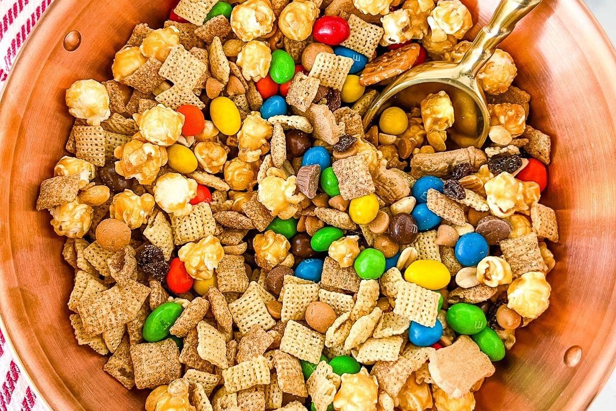 A bowl filled with a festive mix of cereal squares, colorful candies, popcorn, and small cookies offers delightful Christmas snack ideas, with a golden spoon resting on top.