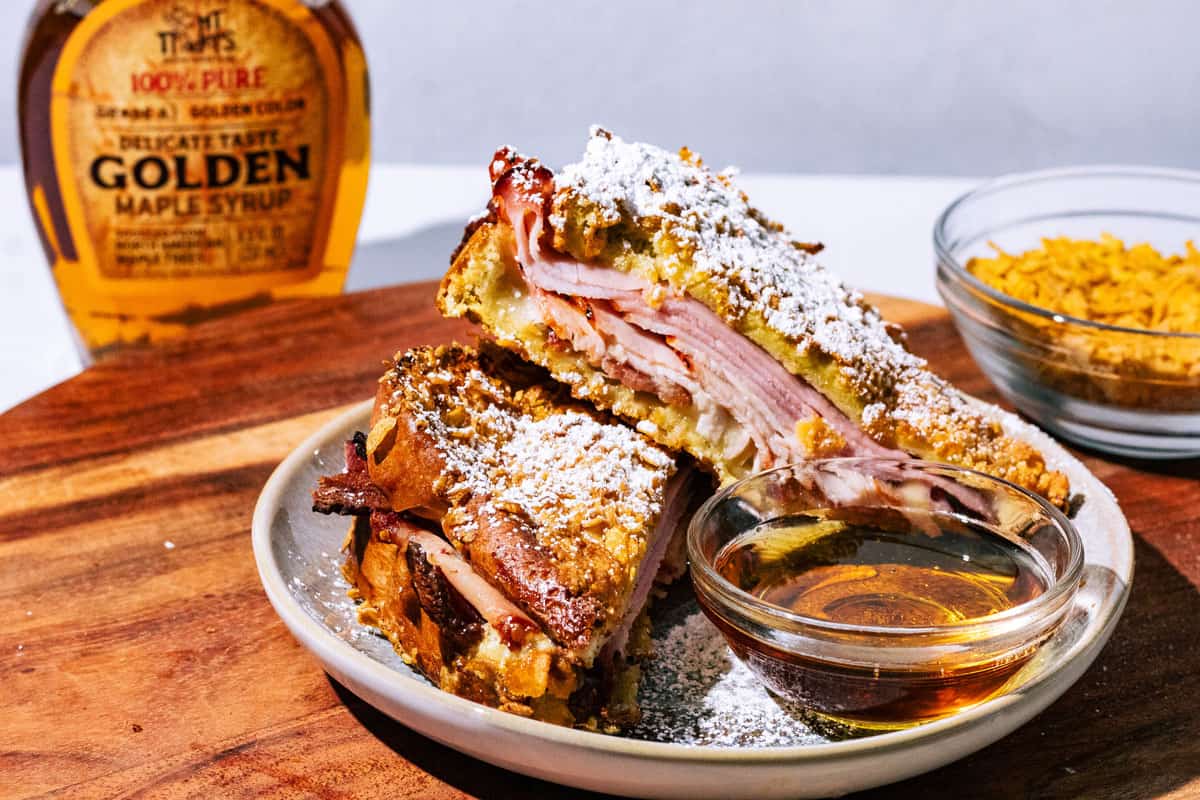 A plate with a Monte Cristo sandwich, sliced in half, dusted with powdered sugar. A small bowl of syrup and a bottle of maple syrup are placed nearby.