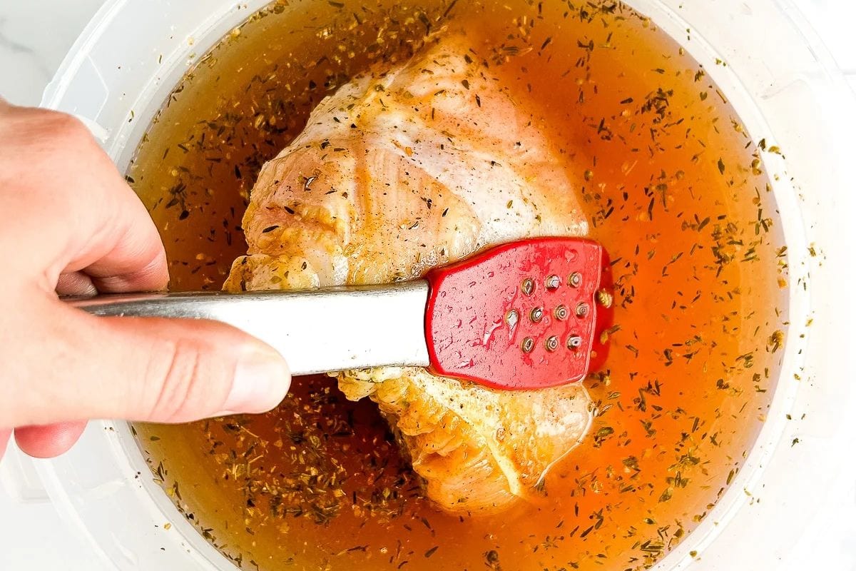 A hand holding red tongs submerges a seasoned turkey breast in a simple, clear brine solution with herbs, inspired by easy turkey brine recipes, in a container.