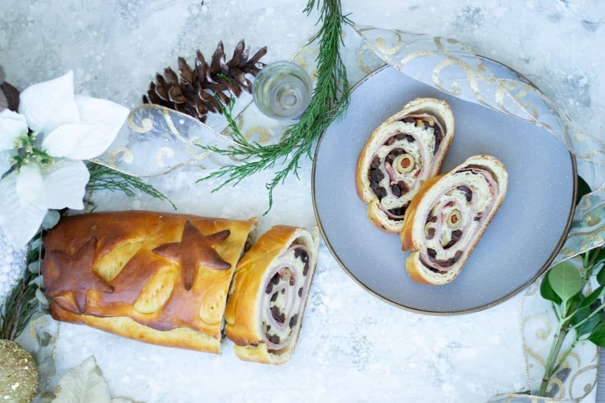 A festive holiday bread roll with filling sits on a plate, surrounded by charming decorations like ribbon, pine cones, and flowers on a light textured background. Perfect for adding a touch of joy to your holiday bread recipes collection.