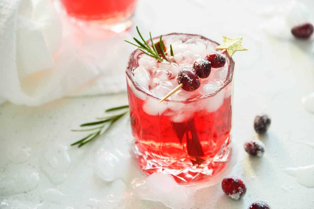 A glass of cranberry cocktail with ice, garnished with a rosemary sprig and skewered cranberries, embodies the essence of holiday cocktail recipes. Additional cranberries and rosemary are artfully scattered around, inviting a festive spirit.