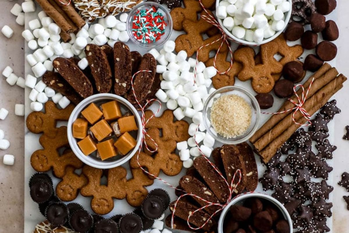 A variety of sweet treats including gingerbread, biscotti, marshmallows, caramel squares, chocolate-covered items, and sprinkles arranged on a board.