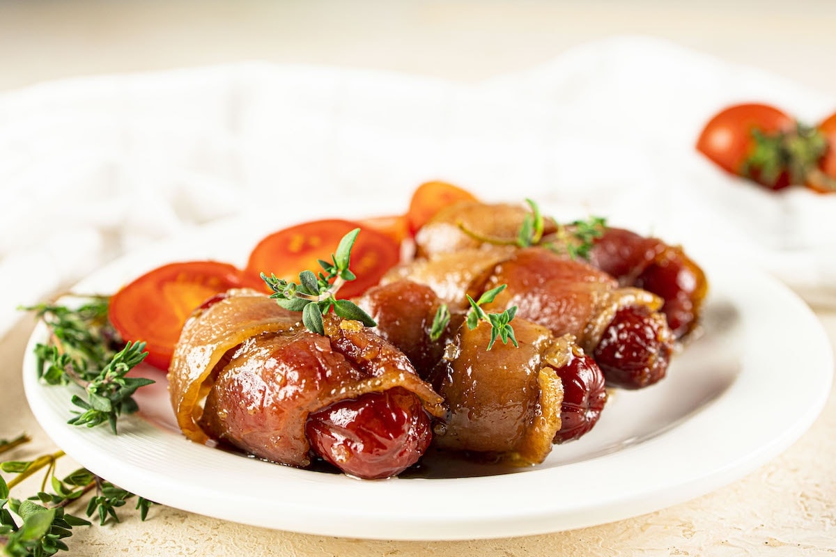 Plate of bacon-wrapped cherry tomatoes garnished with thyme, alongside sliced cherry tomatoes on a white plate.