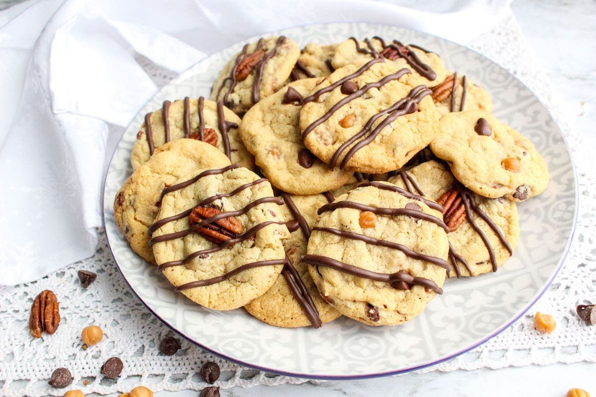 A plate of chocolate-drizzled cookies with pecans and chocolate chips on a patterned dish.