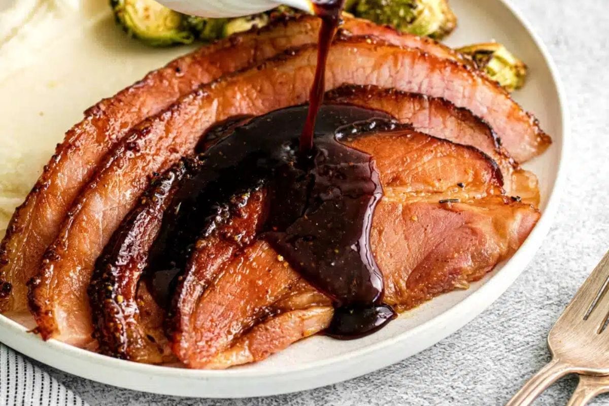 Sliced glazed ham on a plate with a dark sauce being poured over it, accompanied by Brussels sprouts—a delightful addition to any Thanksgiving food spread.