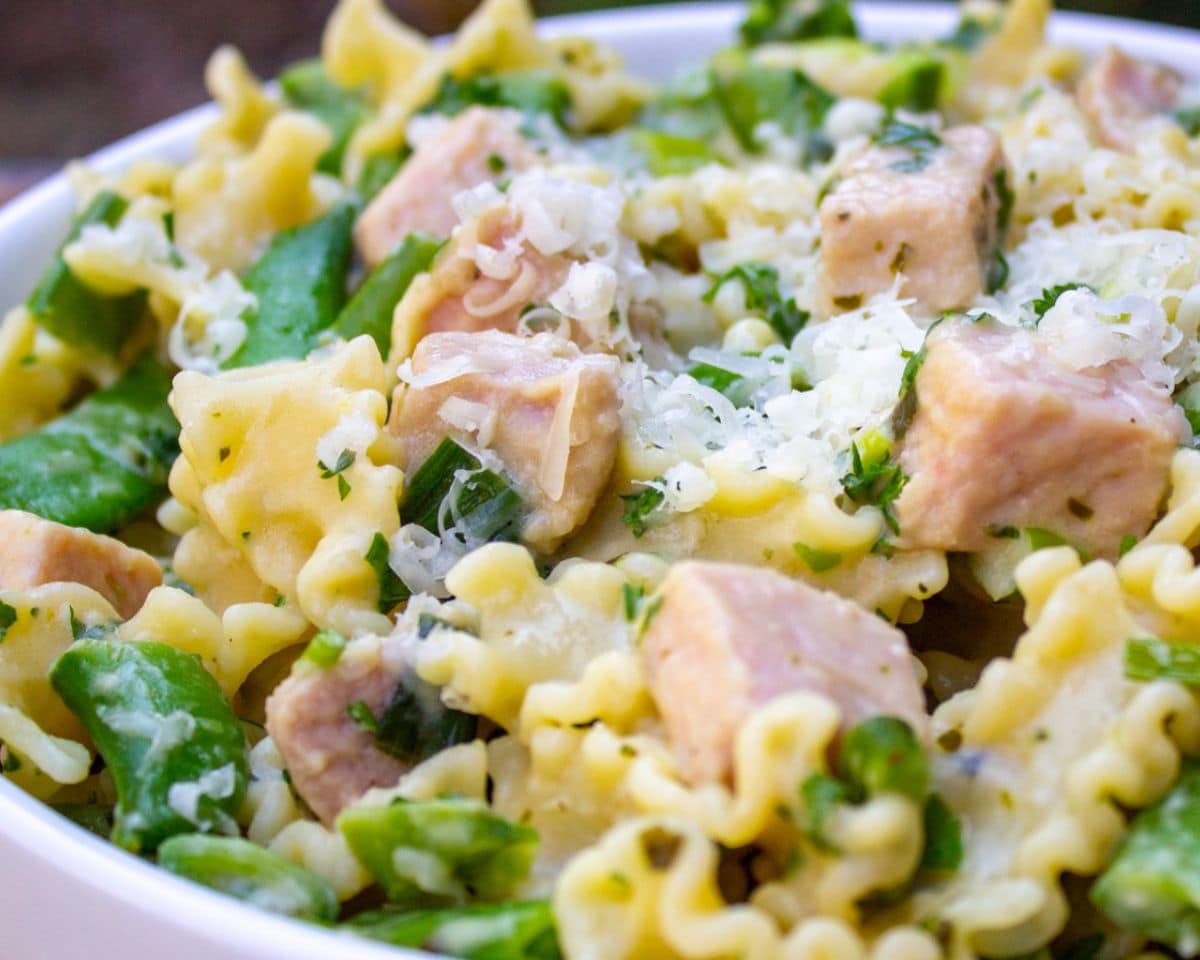 A close-up of pasta with chunks of chicken, snap peas, and shredded cheese in a white bowl, perfect for reinventing leftover ham recipes.