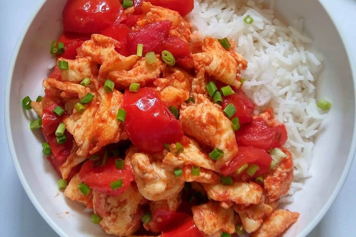 A bowl of rice topped with cooked chicken pieces, tomatoes, and chopped green onions.